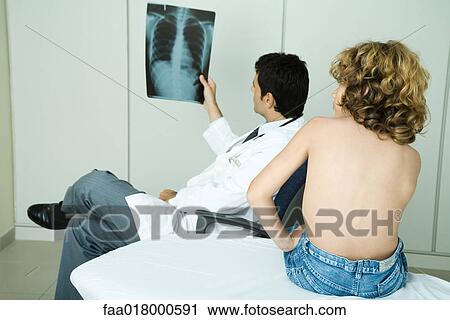 Doctor looking at x-ray while boy sits barechested on examination table View Large Photo Image Stock Image - Doctor looking at x-ray while boy sits barechested on examination table. Fotosearch
