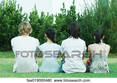Stock Image - Four young friends sitting on grass, side by side, rear view. Fotosearch
