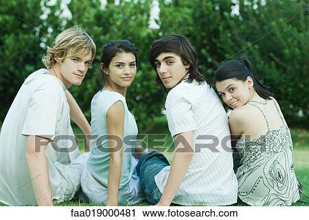 Stock Image - Four young friends sitting on grass, side by side, looking over shoulders at camera. Fotosearch