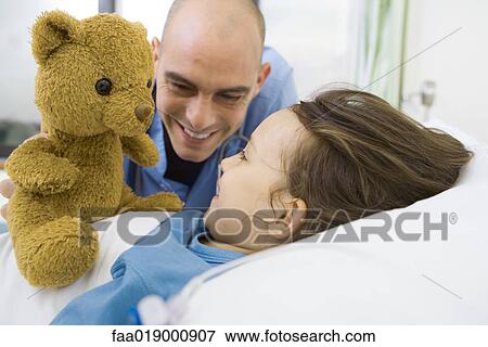 Stock Photo - Girl lying in hospital bed, nurse holding up teddy bear. Fotosearch