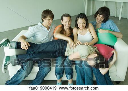 Group of young friends sitting on sofa, one lying across legs of others View Large Photo Image Stock Photo - Group of young friends sitting on sofa, one lying across legs of others. Fotosearch