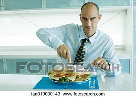 Stock Image - Man cutting into large sandwich with knife and fork. Fotosearch