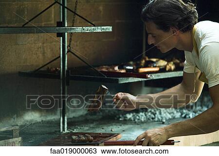 Stock Image - Man grilling meat on barbecue, putting meat onto wooden board. Fotosearch