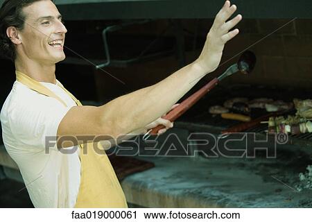Stock Image - Man grilling meat on barbecue, waving out of frame. Fotosearch