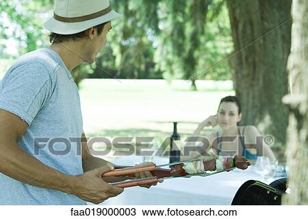 Stock Image - Man holding kebab, turning toward woman sitting at picnic table. Fotosearch