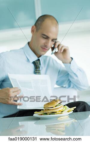Man sitting at table with hamburger, reading newspaper and using cell phone View Large Photo Image Stock Image - Man sitting at table with hamburger, reading newspaper and using cell phone . Fotosearch