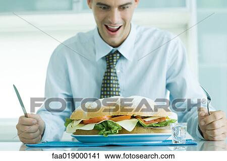 Stock Image - Man sitting holding knife and fork, looking down at large sandwich. Fotosearch