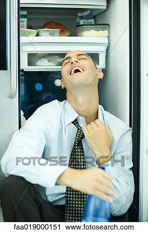 Stock Image - Man sitting in front of open refrigerator, pulling on collar, head back. Fotosearch