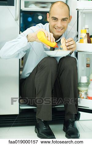 Man sitting in front of open refrigerator, putting mustard on hamburger View Large Photo Image Stock Photo - Man sitting in front of open refrigerator, putting mustard on hamburger. Fotosearch