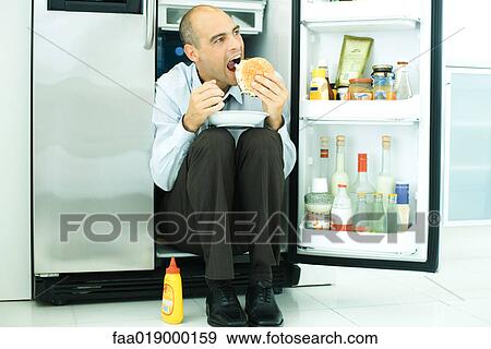 Stock Photo - Man sitting in front of open refrigerator, eating hamburger. Fotosearch