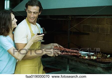 Stock Image - Man tending barbecue, while woman holds up glass of wine for him. Fotosearch