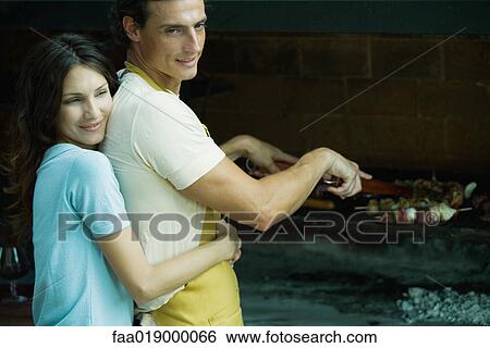Man tending barbecue, woman hugging man from behind View Large Photo Image Stock Photograph - Man tending barbecue, woman hugging man from behind. Fotosearch