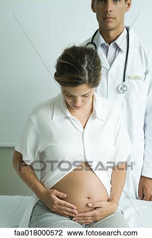 Stock Image - Pregnant woman sitting on examination table, looking down at stomach, doctor standing behind her. Fotosearch