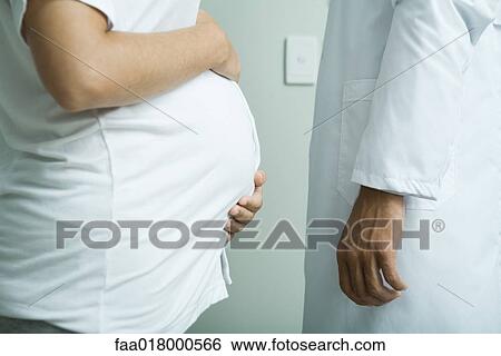 Stock Photograph - Pregnant woman standing face to face with doctor, mid section, side view. Fotosearch
