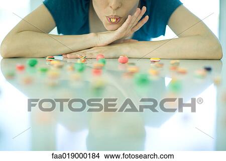 Picture - Table spread with candy, woman resting head on arms, holding piece of candy between teeth. Fotosearch