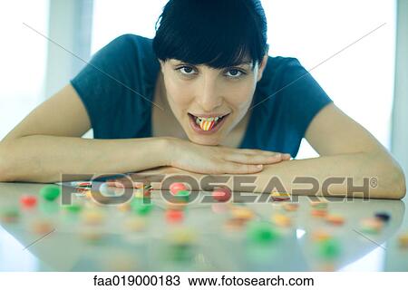Stock Image - Table spread with candy, woman resting head on arms, holding piece of candy between teeth. Fotosearch