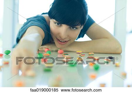 Stock Photography - Table spread with candy, woman resting head on arms, reaching for piece of candy. Fotosearch
