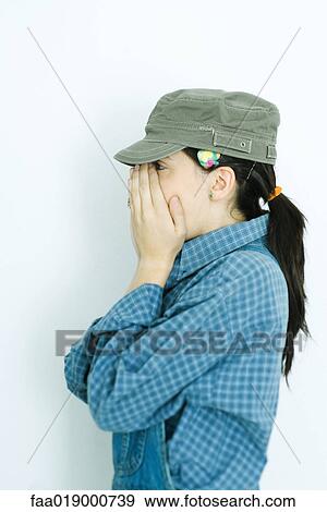 Stock Photo - Teen girl wearing overalls and cap, covering face, side view. Fotosearch