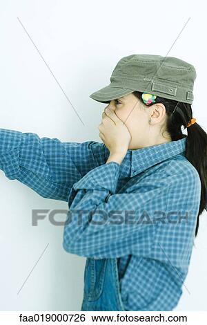 Stock Photograph - Teen girl wearing overalls and cap, covering mouth and pointing. Fotosearch