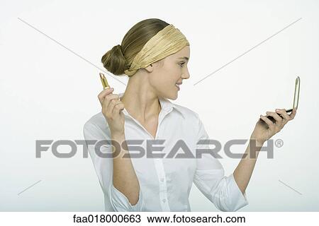 Stock Image - Teenage girl holding lipstick, looking at self in hand mirror. Fotosearch