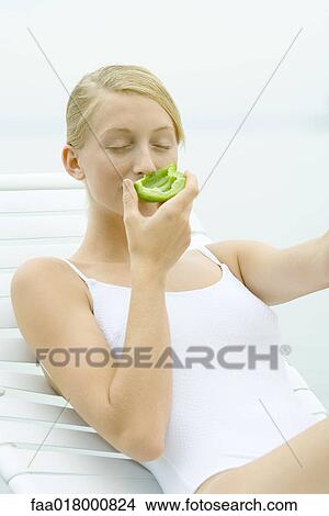 Teenage girl wearing bathing suit, sitting in lounge chair, smelling piece of green pepper View Large Photo Image Picture - Teenage girl wearing bathing suit, sitting in lounge chair, smelling piece of green pepper. Fotosearch