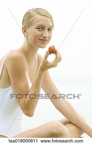 Stock Image - Teenage girl wearing bathing suit, holding up strawberry, smiling. Fotosearch