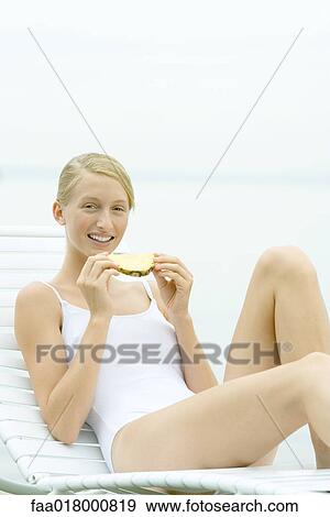 Stock Photo - Teenage girl wearing bathing suit, sitting in lounge chair, eating slice of pineapple. Fotosearch