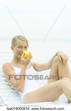 Stock Photograph - Teenage girl wearing bathing suit, sitting in lounge chair, eating apple. Fotosearch