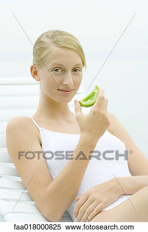 Stock Photography - Teenage girl wearing bathing suit, sitting in lounge chair, holding piece of green pepper. Fotosearch