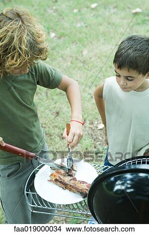 Two boys standing next to barbecue, one cutting up meat on plate View Large Photo Image Picture - Two boys standing next to barbecue, one cutting up meat on plate. Fotosearch