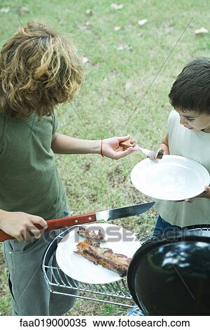 Stock Photography - Two boys standing next to barbecue, one putting piece of meat onto other boy's plate. Fotosearch