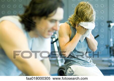 Stock Photography - Two men riding exercise bikes, one wiping face. Fotosearch