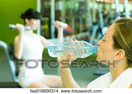 Picture - Two women in weight room, focus on woman in foreground drinking bottle of water. Fotosearch