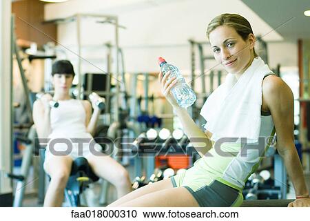 Stock Image - Two women in weight room, focus on woman holding bottle of water. Fotosearch