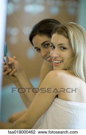 Stock Image - Two women wrapped in towels, holding up cell phone together, close-up. Fotosearch