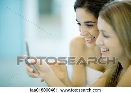 Stock Image - Two women wrapped in towels, looking at cell phone together, close-up. Fotosearch