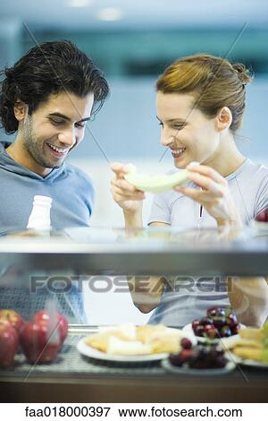 Stock Photo - Two young adults standing, having healthy snack in cafeteria. Fotosearch