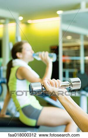 Stock Image - Woman drinking bottle of water, focus on second woman's arm holding dumbbell in foreground. Fotosearch