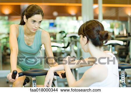 Picture - Woman riding exercise bike, talking to second woman. Fotosearch