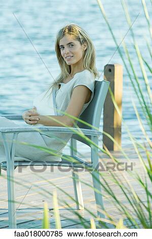 Stock Photography - Woman sitting on dock with book on lap, looking at camera. Fotosearch