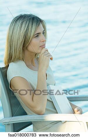 Stock Image - Woman sitting with book on lap, looking toward distance. Fotosearch