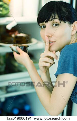 Stock Image - Woman taking piece of cake from refrigerator, looking at camera with finger over lips. Fotosearch