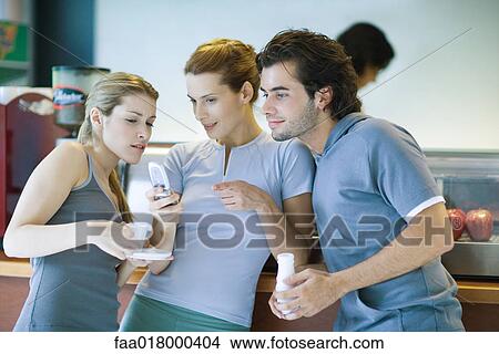 Picture - Young adults dressed in exercise clothes, standing by snack bar, looking at cell phone. Fotosearch
