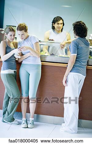 Stock Photography - Young adults dressed in exercise clothes, standing by snack bar, chatting. Fotosearch