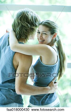 Stock Image - Young couple in exercise clothing standing by window, woman looking over shoulder, smiling at camera. Fotosearch