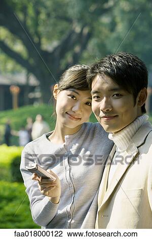 Stock Image - Young couple with cell phone, smiling at camera, portrait. Fotosearch