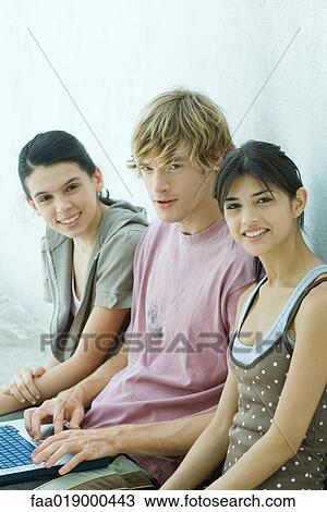 Stock Image - Young friends using laptop together, smiling at camera. Fotosearch