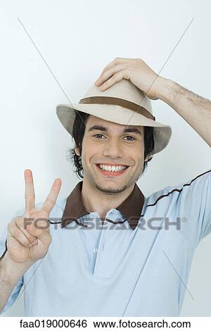 Young man holding hat on head, holding up fingers in peace sign View Large Photo Image Stock Photograph - Young man holding hat on head, holding up fingers in peace sign. Fotosearch