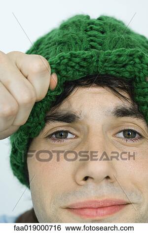 Young man lifting up edge of knit hat, looking at camera, close-up View Large Photo Image Stock Photograph - Young man lifting up edge of knit hat, looking at camera, close-up. Fotosearch