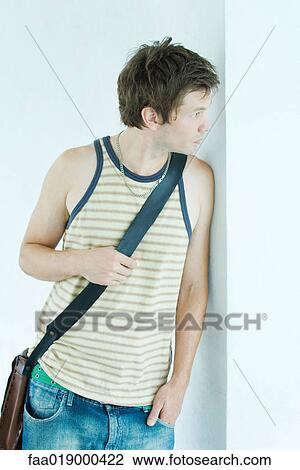 Young man wearing messenger bag, leaning against wall, looking away View Large Photo Image Stock Image - Young man wearing messenger bag, leaning against wall, looking away. Fotosearch